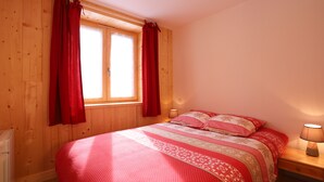 Une chambre avec un plancher en bois, un lit recouvert d’une literie à motifs, une table de chevet avec une lampe et une fenêtre ornée de rideaux rouges.
