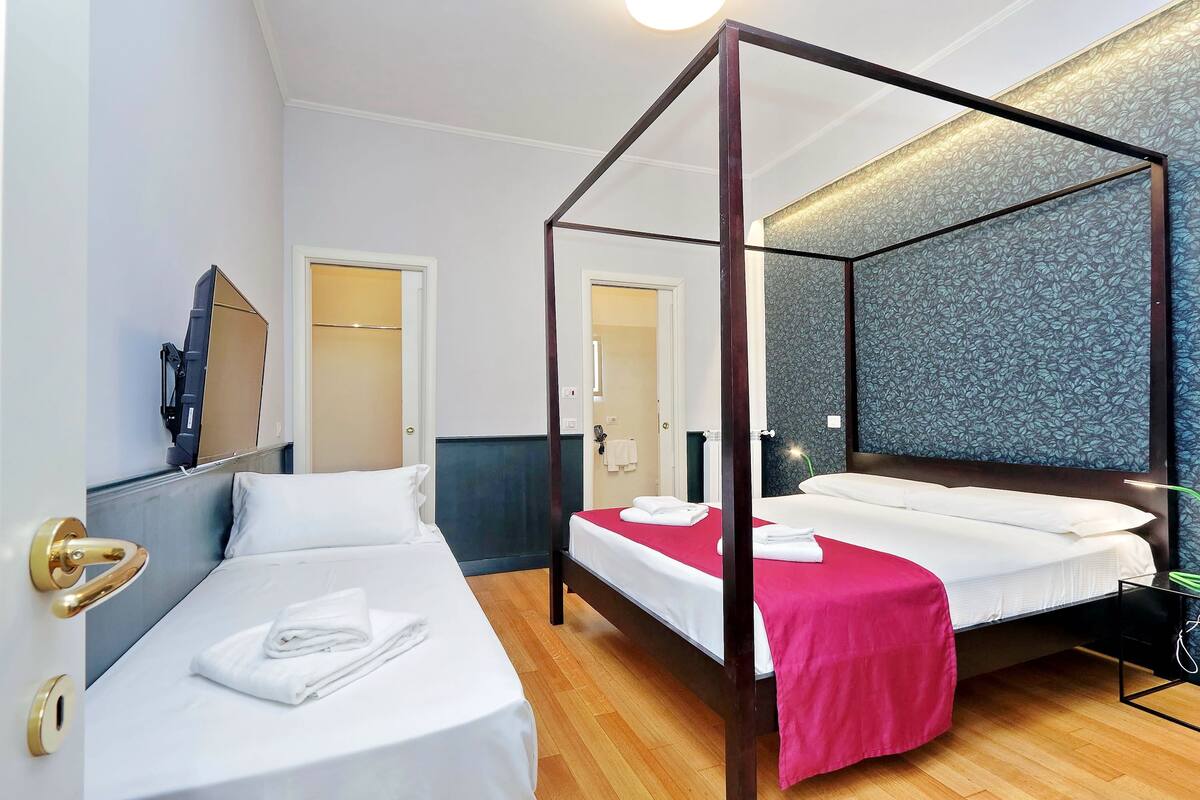 A room with two single beds, a four-poster bed, wooden flooring, a mirror, and a patterned wall.