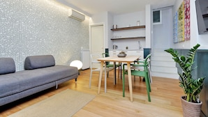 Un salon moderne avec un canapé gris, une table à manger en bois et des chaises, un petit coin cuisine et une plante en pot.
