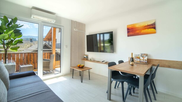 A modern living room with a sofa, dining table, chairs, a TV, and a balcony with a view of a building and trees.