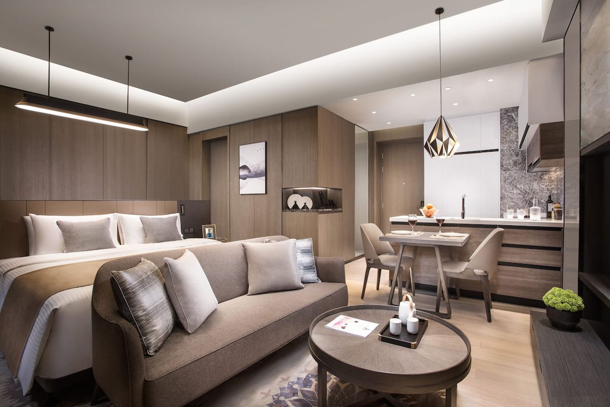 A modern hotel room with a large bed, a sofa, a dining area, and a kitchen.