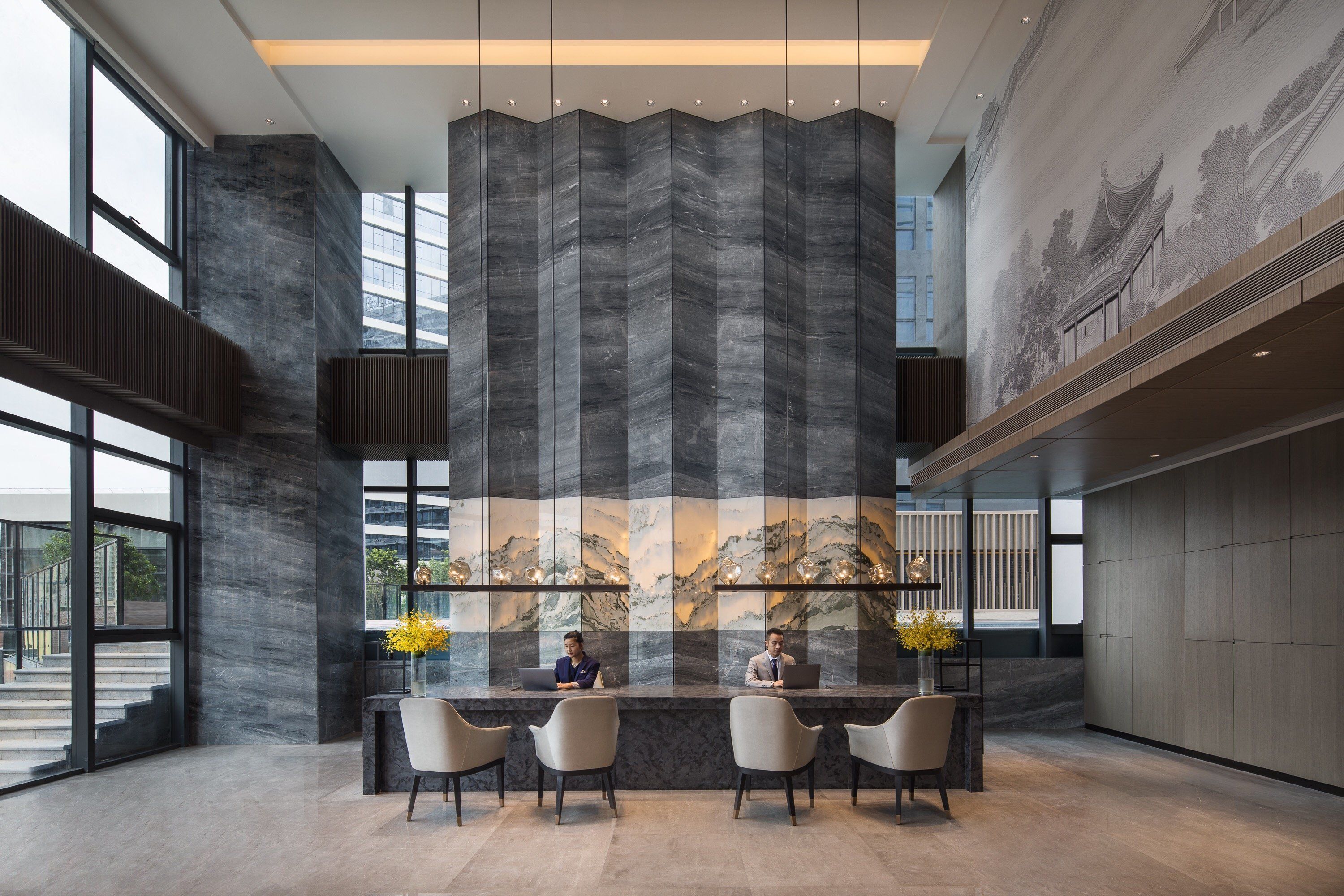 A modern reception area with a large, textured wall and a mural depicting a traditional scene. There are three receptionists seated at a counter with chairs and a vase of yellow flowers.