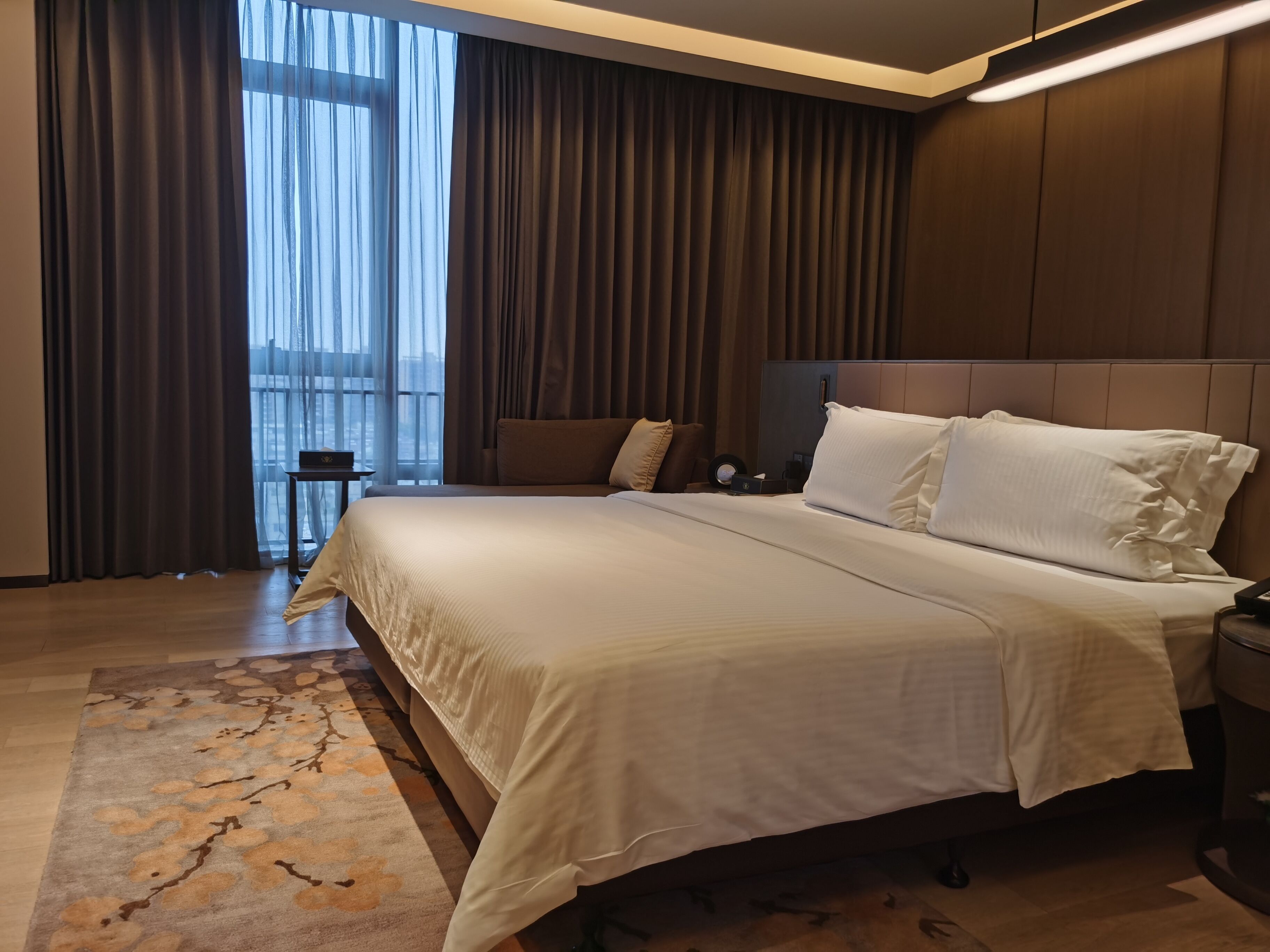A modern hotel room with a large bed, a nightstand, and a view of the city through sheer curtains.