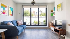 Ett modernt vardagsrum med en blå soffa, en tv på ett träställ och ett stort fönster med utsikt över grönska och byggnader.