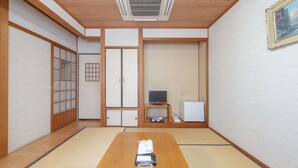 Ein traditioneller japanischer Raum mit Tatami-Böden, einem Holztisch mit einem Glas und einem Buch, einem Fernseher auf einem flachen Schrank und einem kleinen Kühlschrank.