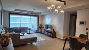 A modern living room with a sofa, coffee table, and dining area. There is a TV on a stand and a plant in the corner. - Marina Casablanca by Saloua (Casablanca)