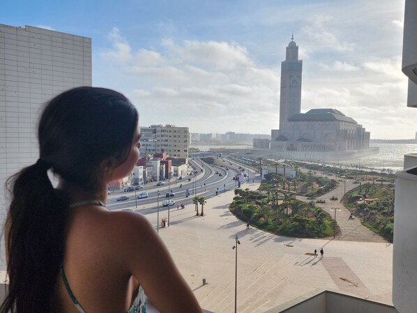 Vista de un paisaje urbano con una torre destacada, una calle concurrida y un paseo marítimo con zonas verdes.