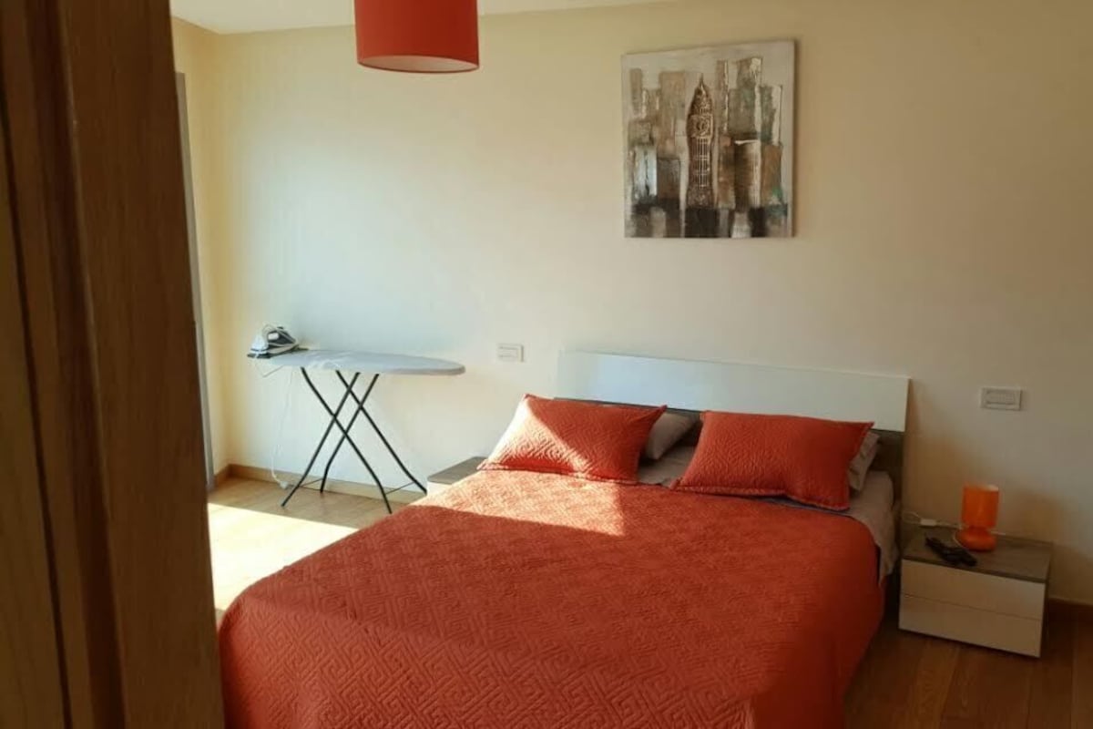 Un dormitorio con una cama, una mesita de noche, una tabla de planchar y un cuadro en la pared.