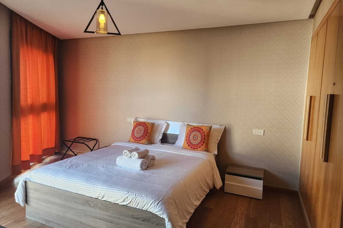 Un dormitorio con una cama, dos almohadas, una mesita de noche y una lámpara colgante.