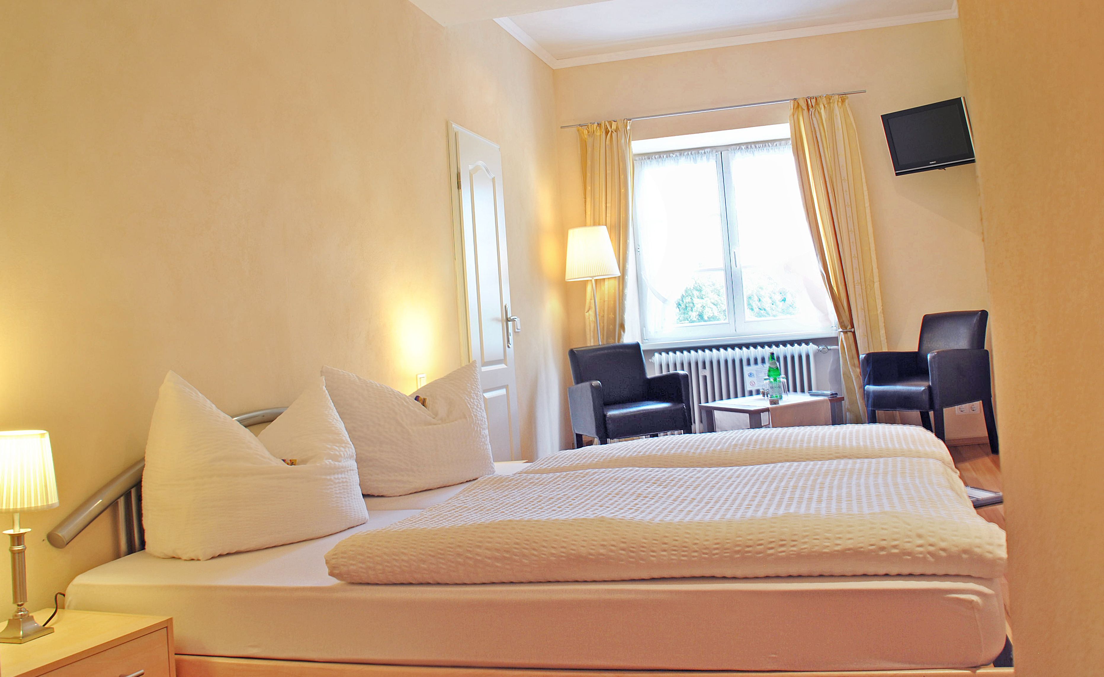 A hotel room with a bed, bedside tables, a desk, two chairs, a lamp, and a TV on the wall.