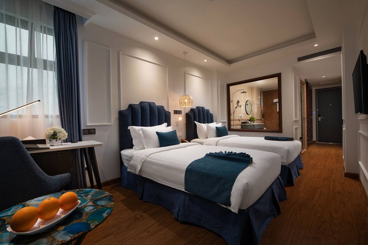 A hotel room with two beds, a desk with a chair, a bedside table with a vase of flowers, and a wooden floor.