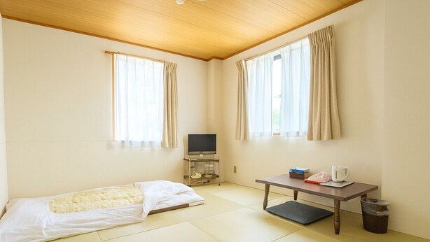 Una habitación minimalista con suelo de tatami, una mesita con un hervidor, un televisor en un soporte y una cama estilo futón.