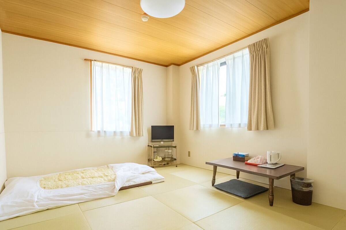 Una habitación minimalista con suelo de tatami, una mesita con un hervidor, un televisor en un soporte y una cama estilo futón.