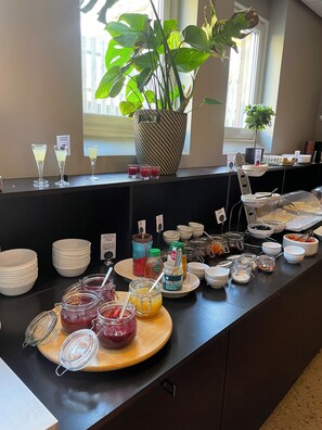 A buffet setup with various food and drink options, including jars of preserves, a large potted plant, and a window with natural light. -  Lidingö Arena Hotel (Lidingo)