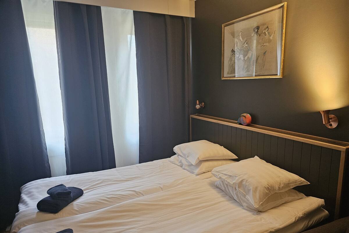 A hotel room with two beds, a framed artwork on the wall, a shelf with a decorative item, and a window with curtains.
