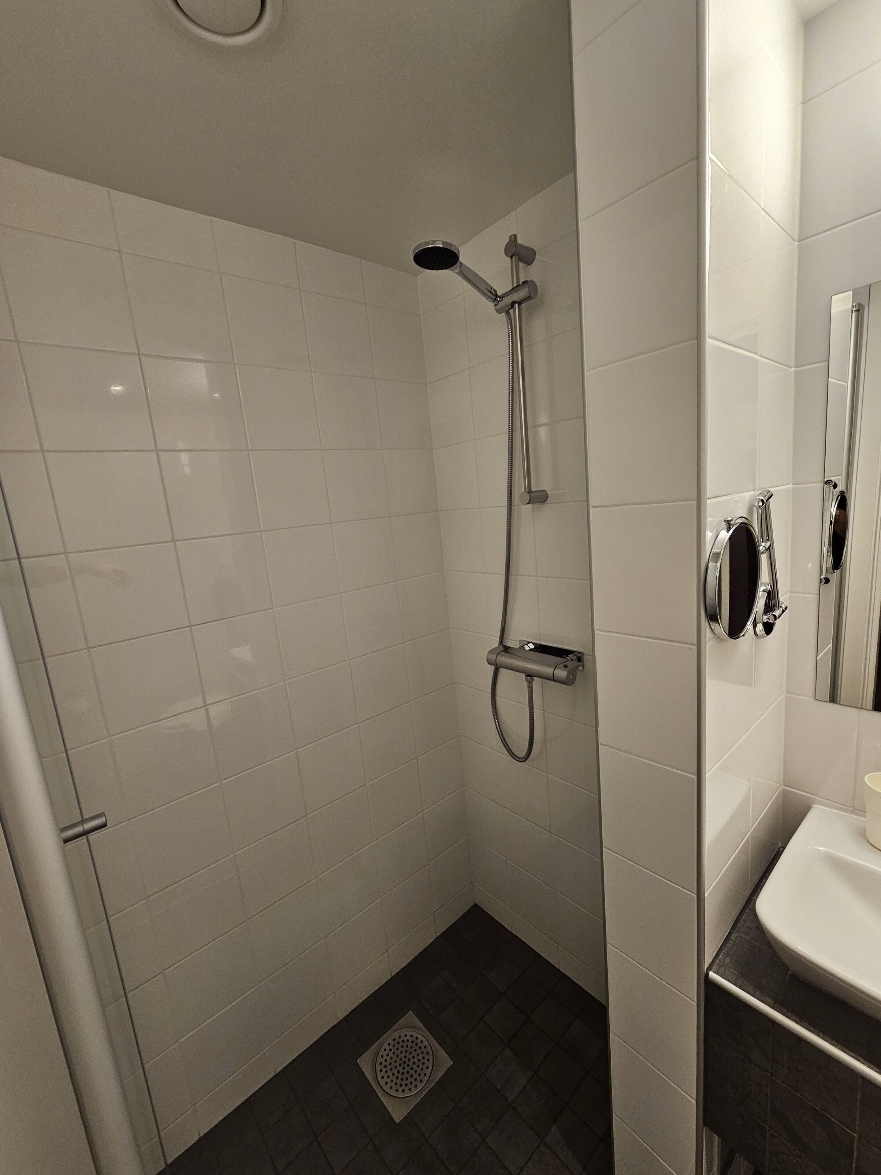 A bathroom with white tiles, a shower with a handheld showerhead, a toilet, and a sink with a mirror.