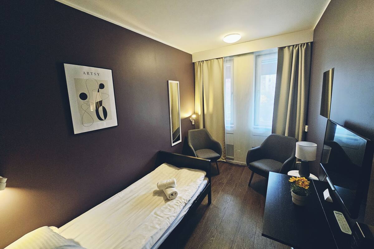 A hotel room with a bed, two armchairs, a desk with a lamp, a TV, and a window with curtains.