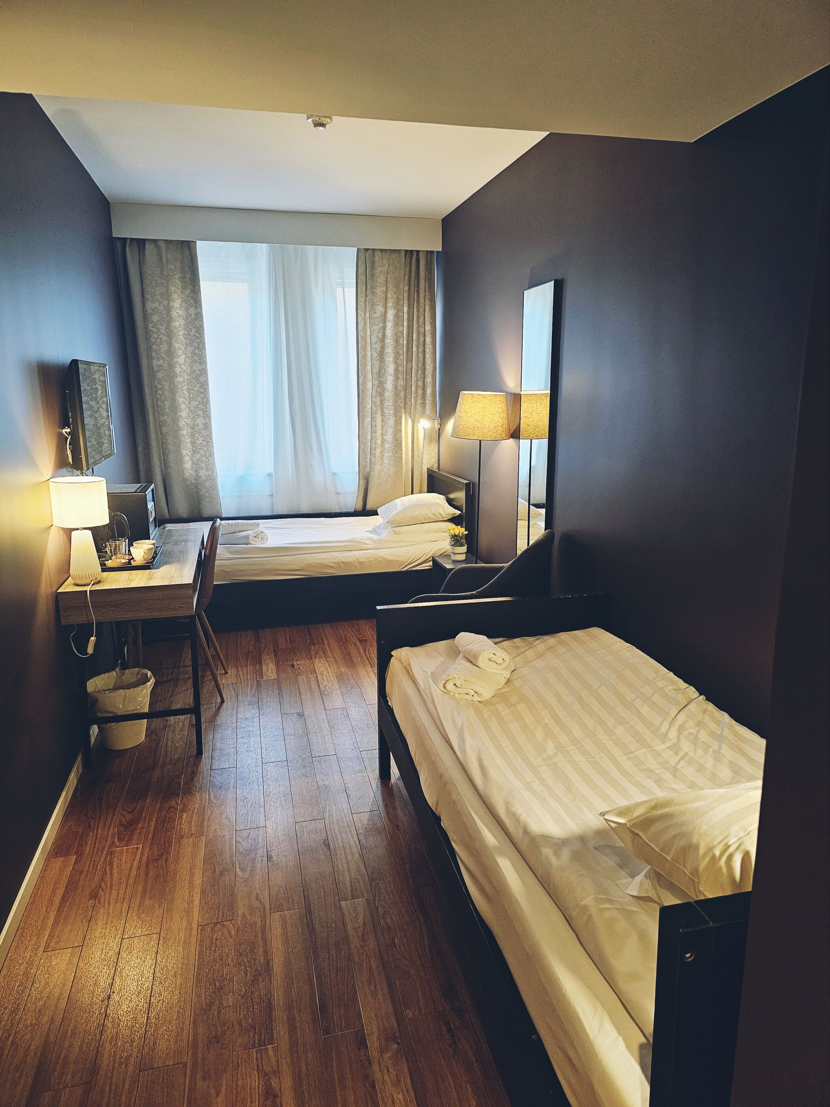 A compact hotel room with a wooden floor, a desk with a lamp, a bed with a rolled towel, a bedside table, and a large mirror.