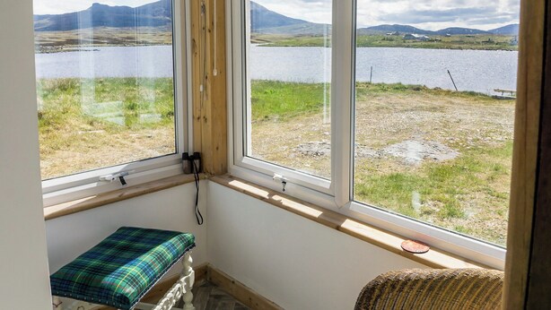 Une pièce avec un banc et une chaise en osier près d’une fenêtre donnant sur un lac et des montagnes.