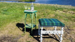 Une table verte avec une bouteille et des verres, un banc à carreaux et un lac avec des montagnes en arrière-plan.