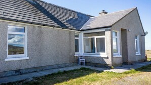 Une maison à deux étages, avec un toit recouvert de tuiles grises, un petit porche avec une chaise blanche et un ciel d’un bleu limpide.