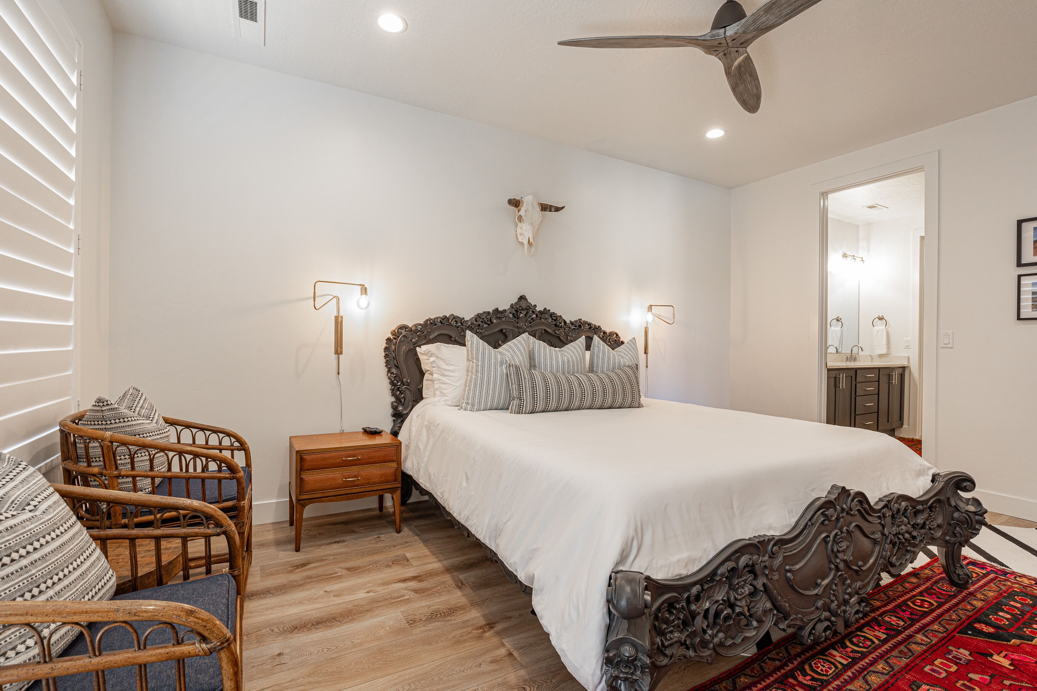 A bedroom with a large bed, wooden nightstands, wicker chairs, a ceiling fan, and a red patterned rug.