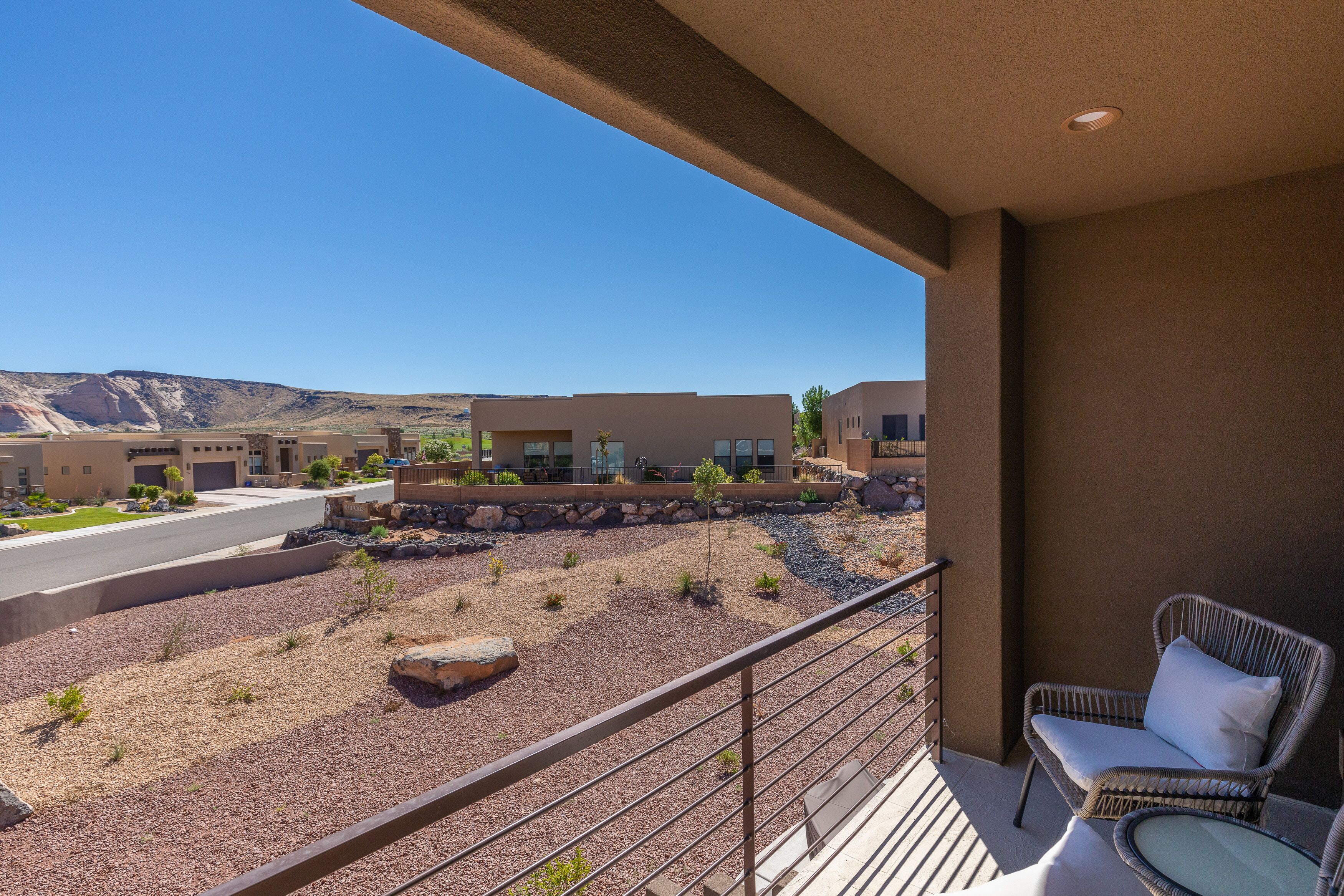 A balcony with a wicker chair and a small table, overlooking a landscaped area with a building in the background.