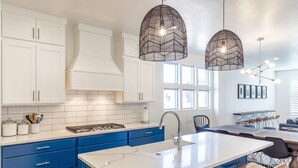 A modern kitchen with white cabinets, blue drawers, a white island with a sink, and pendant lights. - Stunning 4BR Sleeps 18 - Bold Designer Interiors (St. George)
