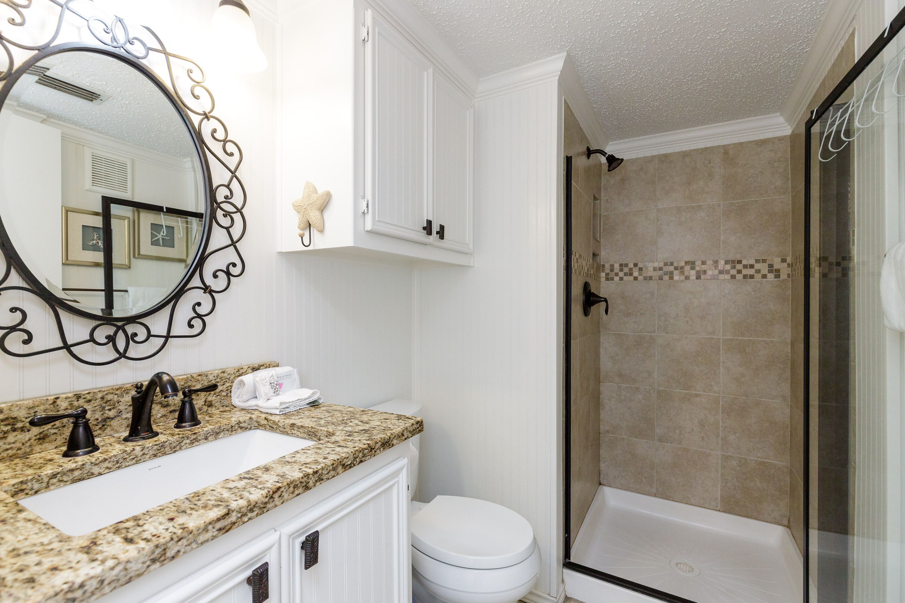 Un baño con lavamanos de encimera de granito, inodoro blanco, ducha con cerramiento de vidrio y espejo decorativo con marco negro.