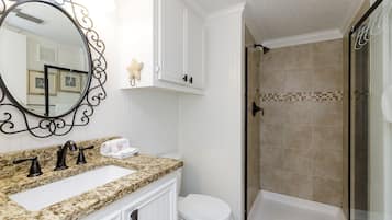 Un baño con lavamanos de encimera de granito, inodoro blanco, ducha con cerramiento de vidrio y espejo decorativo con marco negro.
