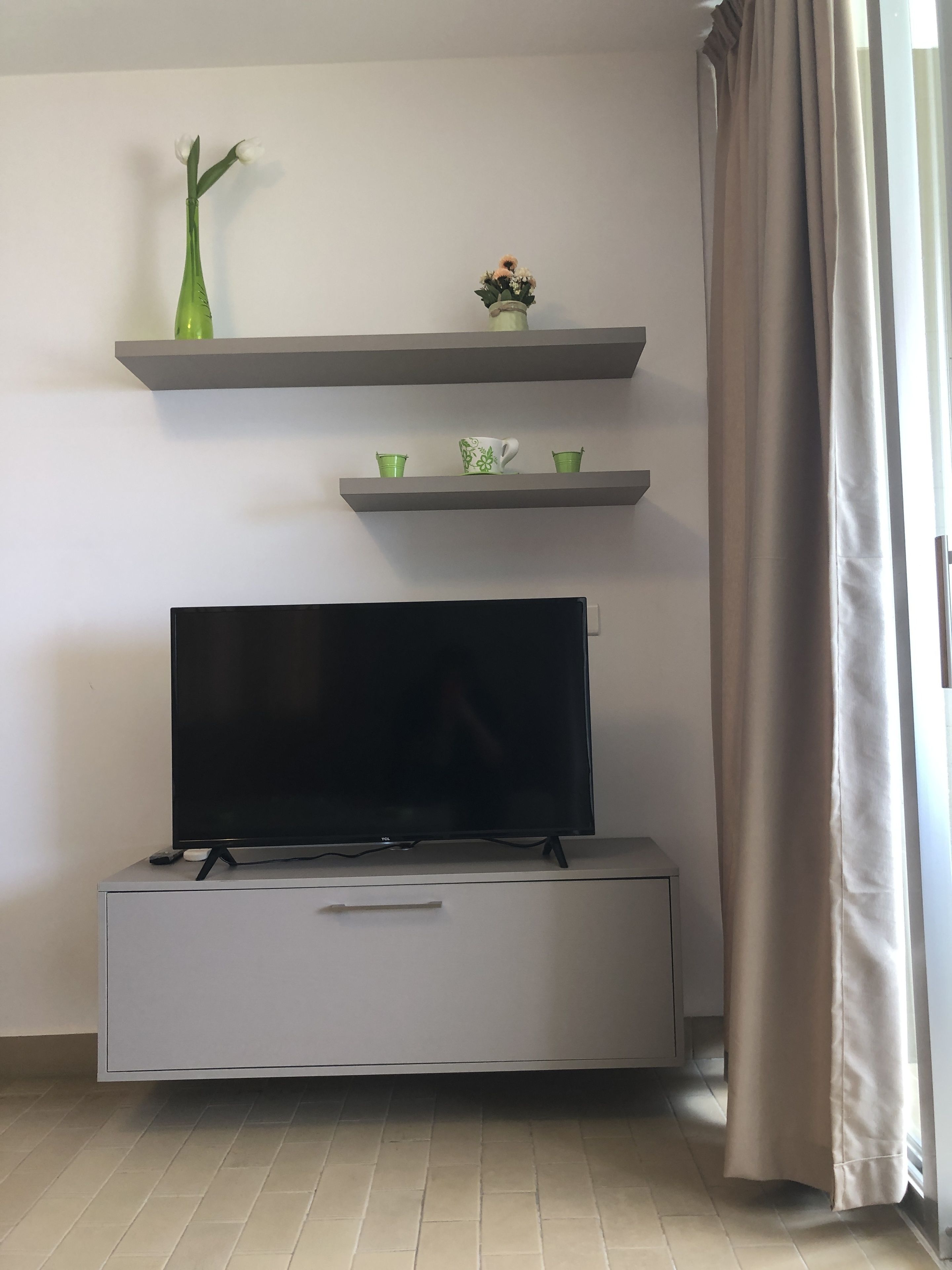 A modern living room with a flat-screen TV on a low cabinet, grey shelves above holding decorative items, and a large window with curtains.