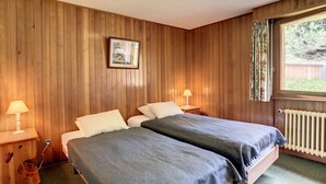 A bedroom with two beds, wooden paneling, a framed picture on the wall, and a window with curtains. - Vouamons A4 (Verbier)
