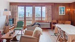 A cozy room with a wooden floor, a green armchair, a beige sofa, a dining table with chairs, a radiator, and a large window with curtains. - Vouamons A4 (Verbier)