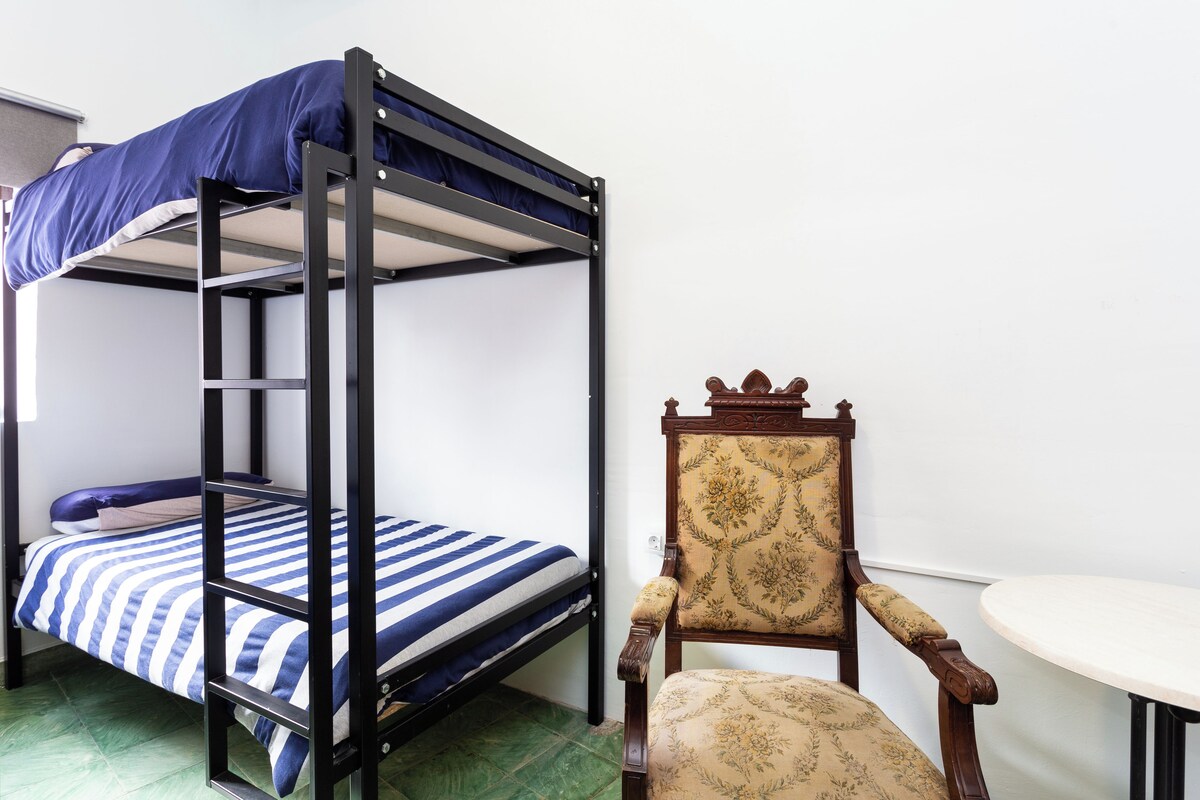 Una cama litera con colcha a rayas azul y blanca, una silla de madera con estampado floral y una mesita redonda.