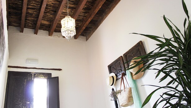 A room with a wooden ceiling, a hanging light fixture, a door, and a wall-mounted hat rack with hats and a basket. - El Hostal Del Cubo (San Juan de la Rambla)
