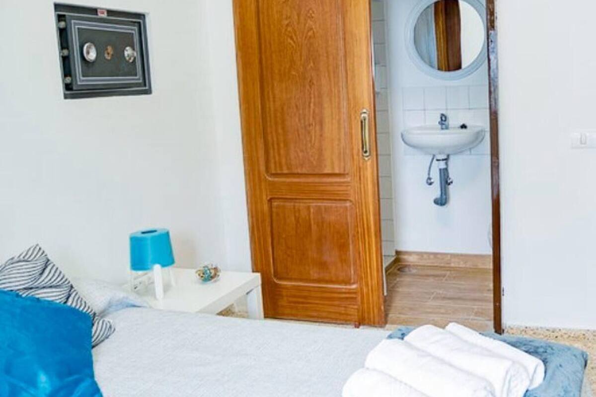 A bedroom with a wooden wardrobe, a bed with blue and white bedding, a bedside table with a blue lamp, and a bathroom with a sink and mirror.
