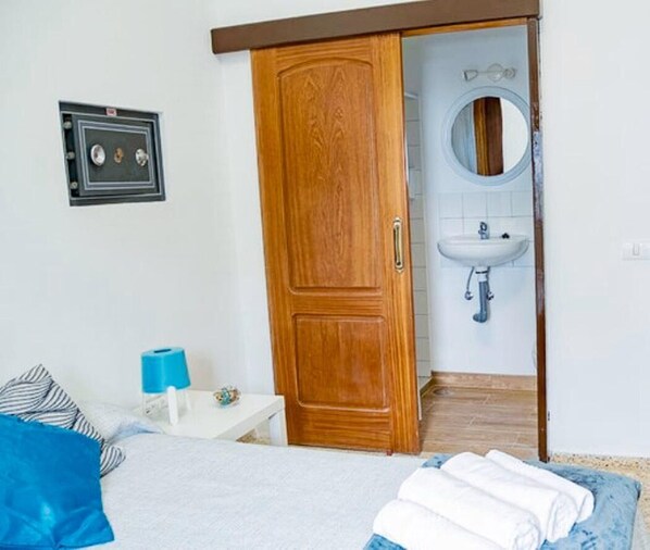 A bedroom with a wooden wardrobe, a bed with blue and white bedding, a bedside table with a blue lamp, and a bathroom with a sink and mirror.