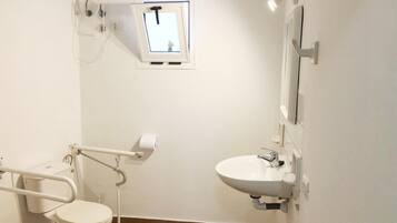 Et badeværelse med en hvid vask, et spejl, et toilet med gelænder og et lille vindue med udsigt til en bygning.