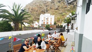 A terrace with wooden tables and chairs, a potted plant, and a view of a white building with a bell tower in the background. - El Hostal Del Cubo (San Juan de la Rambla)