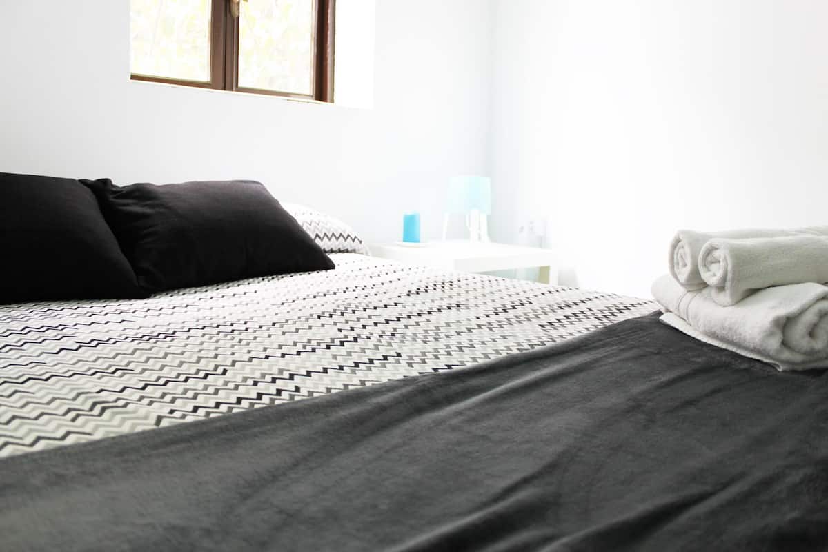 A neatly made bed with a patterned bedspread, a black pillow, and a rolled white towel on the side.