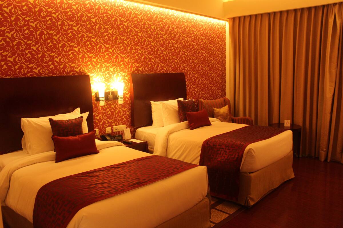 A hotel room with two beds, each with a headboard, white linens, and red throw pillows. There is a nightstand with a lamp and a telephone. The room has a patterned wallpaper and a curtain on the window.