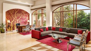 A spacious living area with a large sectional sofa, a glass coffee table, and a red rug. There is a desk with a chair and a lamp in the background. - Ramada by Wyndham Jaipur Jaisinghpura (Jaipur)