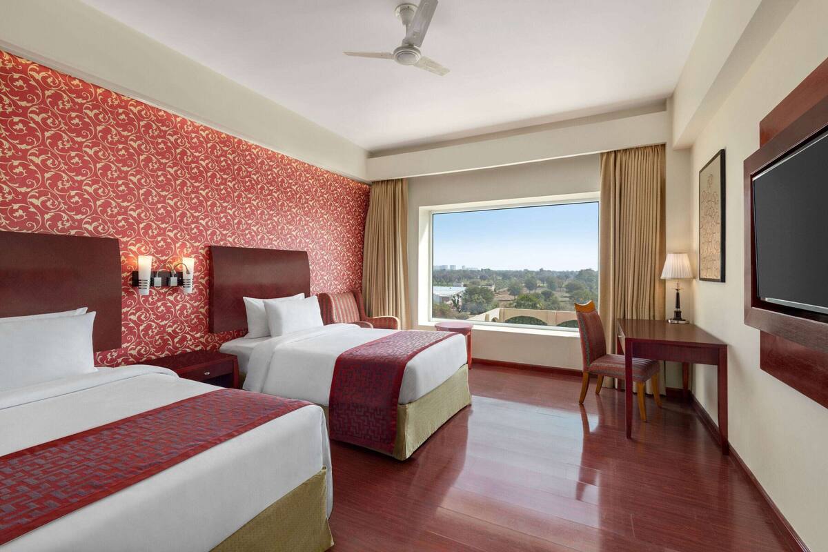 A hotel room with two beds, a desk, a chair, a TV, and a large window with a city view.