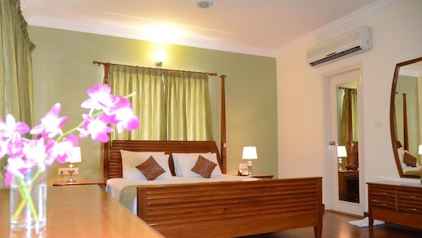 A neatly arranged hotel room with a wooden bed, a bedside table, a mirror, and a green curtain. - Hotel Trinity Suites (Bengaluru)