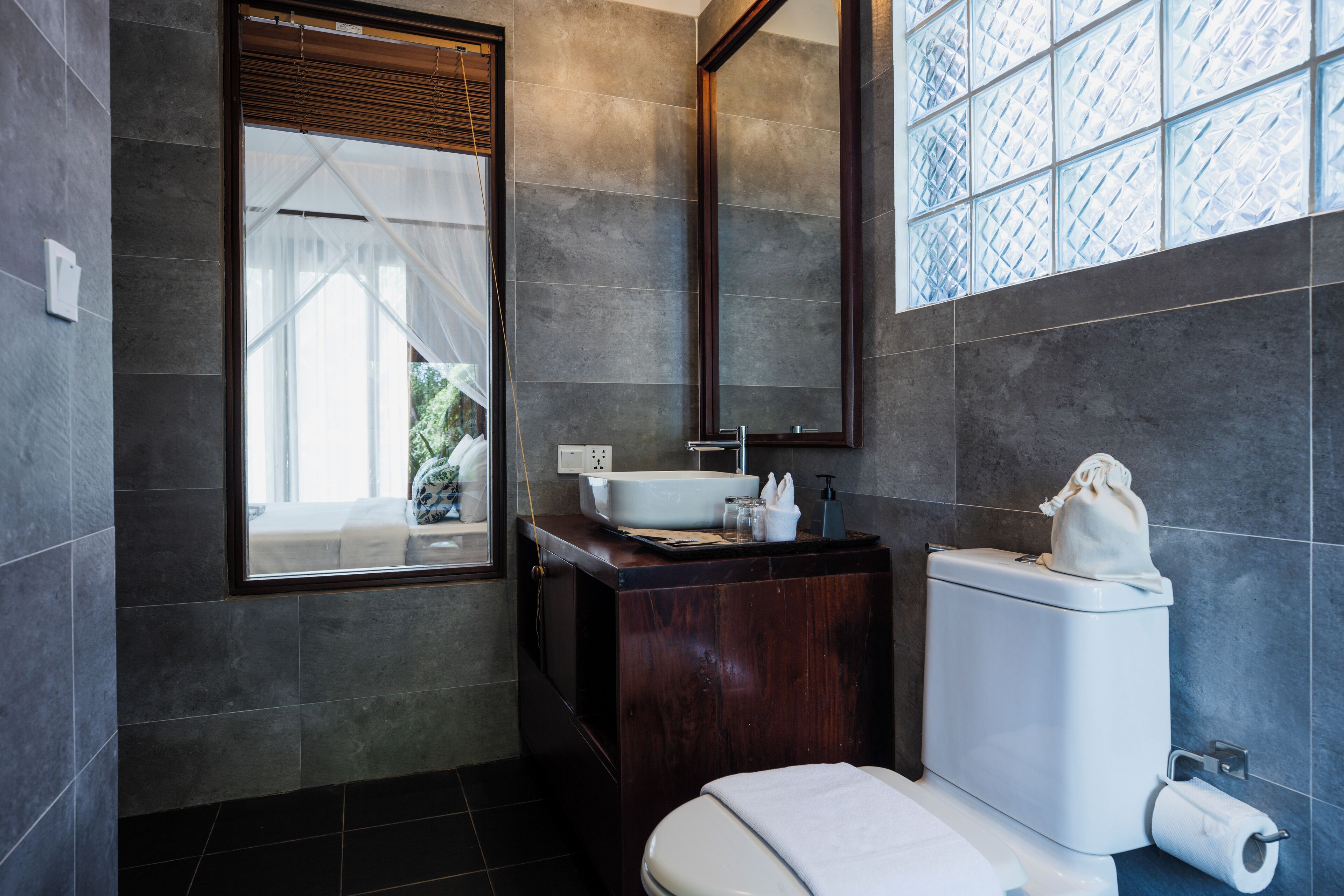 A modern bathroom with a large window, a wooden vanity, a white toilet, and a mirror above the sink.