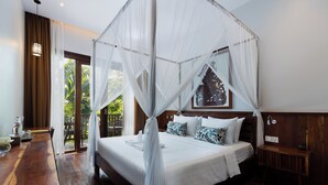 A bedroom with a canopy bed, wooden flooring, a bedside table with a lamp, and a view of greenery outside. - Golden Vishnu Villa (Siem Reap)