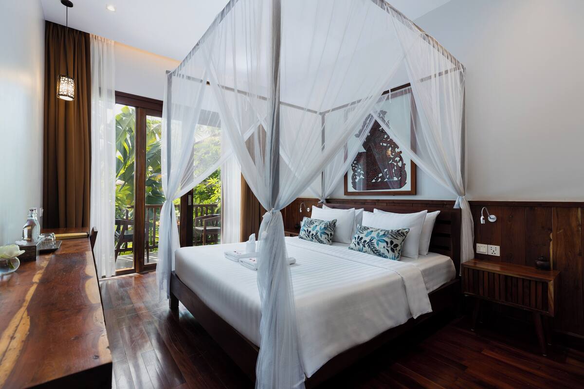 A bedroom with a canopy bed, wooden flooring, a bedside table with a lamp, and a view of greenery outside.