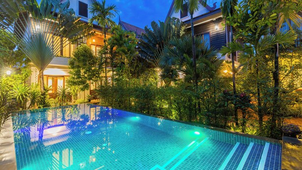 A swimming pool surrounded by lush tropical vegetation and a traditional building with a sloped roof. - Golden Vishnu Villa (Siem Reap)