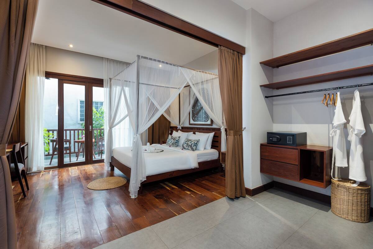 A bedroom with a four-poster bed, a wooden floor, a balcony with outdoor furniture, and a built-in wardrobe.