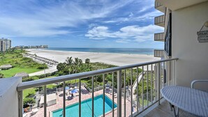 A balcony with a view of a beach, a pool, and a hotel building in the background.